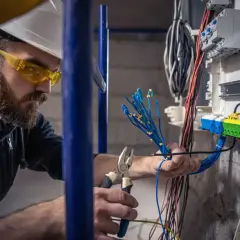 Elektryk z uprawnieniami budowlanymi elektrycznymi pracuje przy instalacji, przygotowując przewody do podłączenia w skrzynce rozdzielczej.