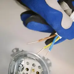 Ręka w rękawicy trzyma przewody elektryczne, przygotowując się do podłączenia fazy w gniazdku. Widoczne są przewody brązowy, niebieski i żółto-zielony.
