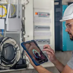Elektryk w kasku i okularach ochronnych wykonuje przegląd instalacji elektrycznej, korzystając z tabletu z aplikacją diagnostyczną.