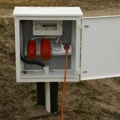 Skrzynka elektryczna na działce bez domu, gotowa do podłączenia prądu. Widoczne bezpieczniki, gniazda i pomarańczowy kabel.