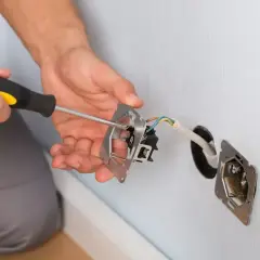 Elektryk wykonuje mostkowanie gniazdka, przykręcając mechanizm do puszki. Widać przewody elektryczne.