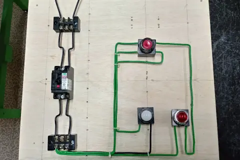 Schemat elektryczny z wyłącznikiem, przyciskami i lampkami. Widoczny jest przewód pe, łączący elementy na drewnianej płycie.