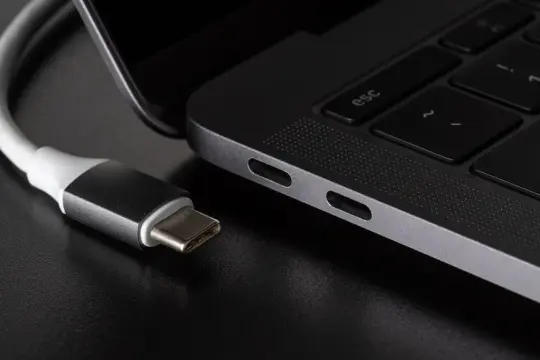 Kabel USB C zbliża się do portu laptopa. Widać jego metalową wtyczkę i biały kabel.