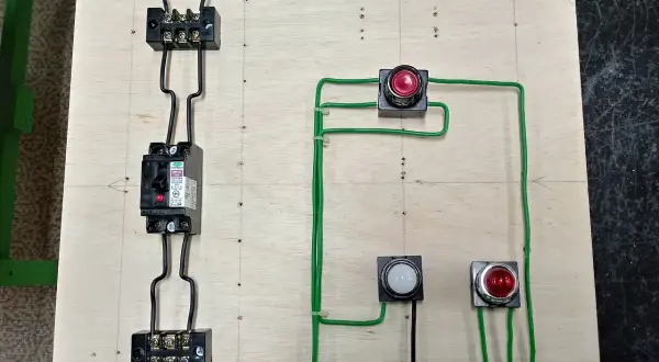 Schemat elektryczny z wyłącznikiem, przyciskami i lampkami. Widoczny jest przewód pe, łączący elementy na drewnianej płycie.