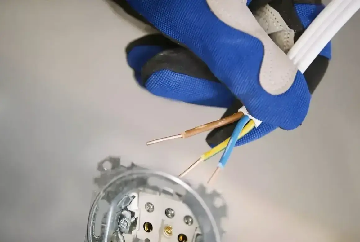 Ręka w rękawicy trzyma przewody elektryczne, przygotowując się do podłączenia fazy w gniazdku. Widoczne są przewody brązowy, niebieski i żółto-zielony.