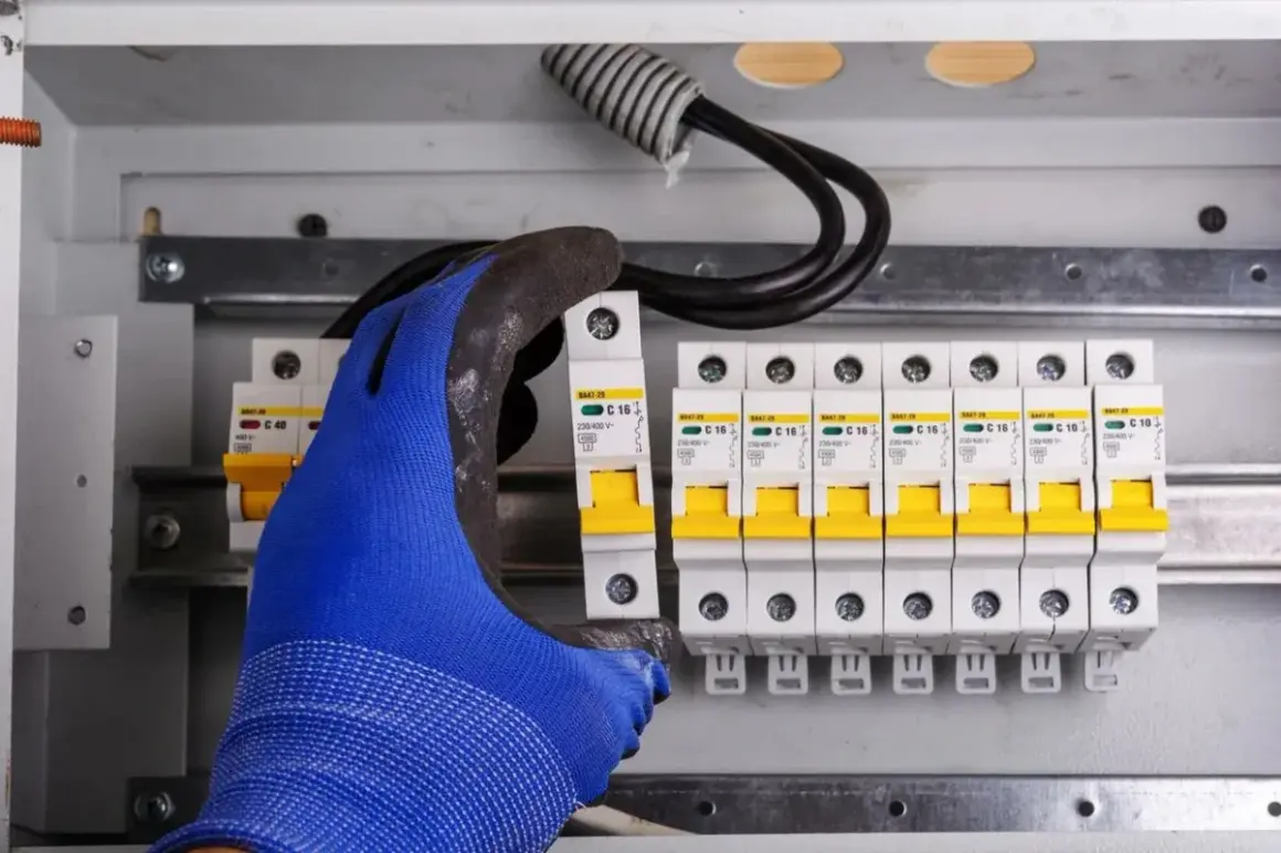 Ręka w niebieskiej rękawicy montuje bezpiecznik w skrzynce. Elektryk zabezpiecza instalację przed przepięciem.
