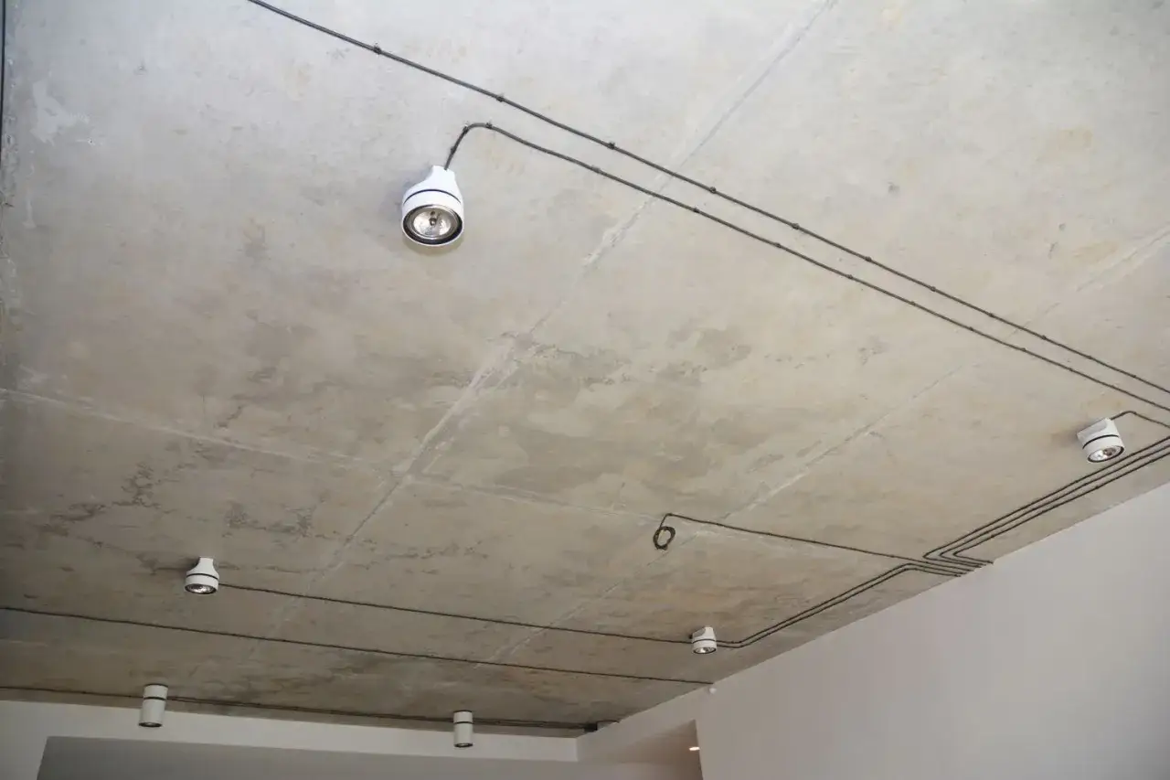Nowoczesna wymiana instalacji elektrycznej bez kucia. Widoczne białe oprawy oświetleniowe na betonowym suficie, połączone czarnymi przewodami.
